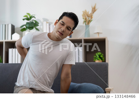 Back Discomfort. Young man experiencing pain while sitting on a couch. Back Discomfort. Young man experiencing pain while sitting on a couch. 133148466