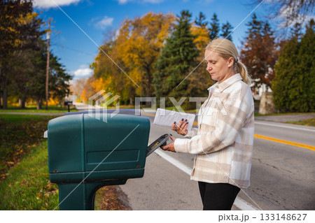 Woman standing by a roadside mailbox placing an envelope inside on a sunny autumn day, with colorful trees and a quiet suburban street in the background. 133148627