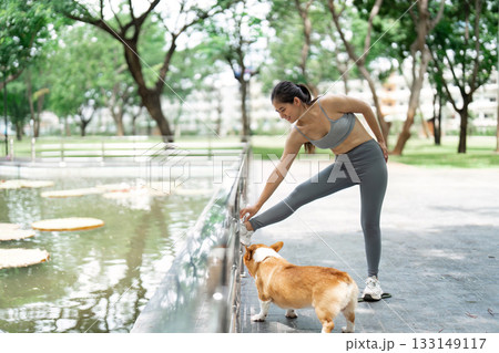 Wellness and Stretching. Young woman stretching with her corgi dog in a serene park. 133149117