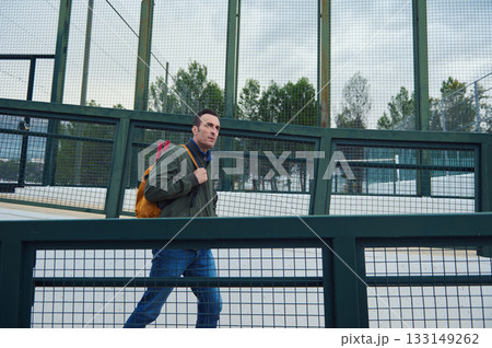 Man With Backpack Walking Through Fenced Sports Court On A Cold Day Man With Backpack Walking Through Fenced Sports Court On A Cold Day 133149262