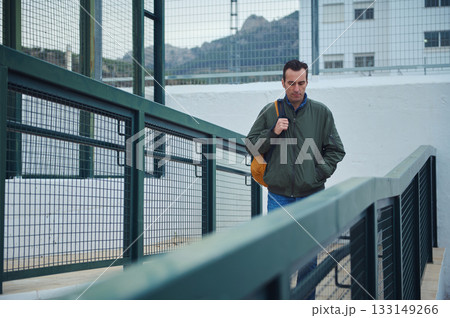 Man With Green Jacket and Orange Backpack Walking Across an Urban Ramp Man With Green Jacket and Orange Backpack Walking Across an Urban Ramp 133149266