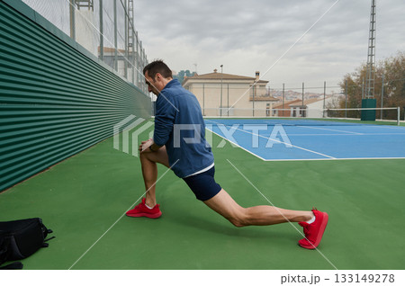 Athletic Man Performs Lunge Stretch On Outdoor Tennis Court Preparing For Workout Session Athletic Man Performs Lunge Stretch On Outdoor Tennis Court Preparing For Workout Session 133149278