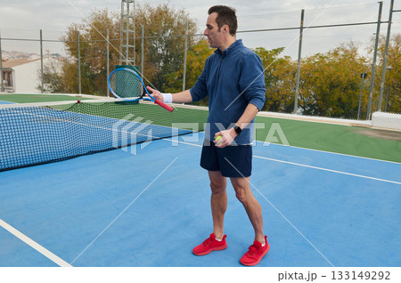 Man In Blue Hoodie Plays Tennis On Outdoor Court With Ball And Racket Man In Blue Hoodie Plays Tennis On Outdoor Court With Ball And Racket 133149292