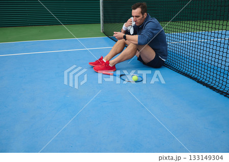 Man Resting After Tennis Practice On Blue Court By Net With Ball And Racket 133149304