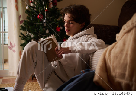Teen Boy Reading a Book by Christmas Tree in a Cozy Home During Holidays Teen Boy Reading a Book by Christmas Tree in a Cozy Home During Holidays 133149470