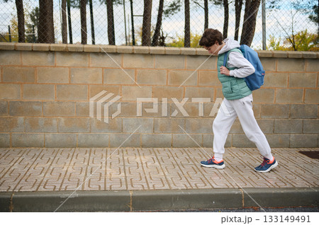 Teen Boy Walking With Backpack On Sidewalk In Casual Winter Outfit Near A Brick Wall Teen Boy Walking With Backpack On Sidewalk In Casual Winter Outfit Near A Brick Wall 133149491