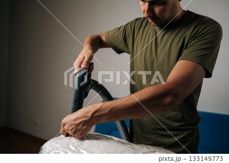 Young man using vacuum cleaner to extract air from storage bag filled with bedding, positioned on ironing board, creating efficient space-saving storage solution. Concept of household chores. 133149773