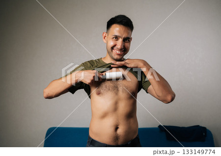 Portrait of happy excited young man lifting shirt and using sticky lint roller to remove hair and fluff from chest at home, showcasing humorous approach to personal grooming, smiling looking camera. 133149774
