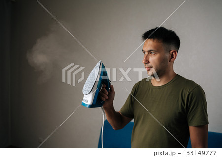Portrait of handsome young man holding modern iron while looking at cloud of steam rising as smoothly presses wrinkles from clothes in tidy domestic setting. Concept of household chores Portrait of handsome young man holding modern iron while looking at cloud of steam rising as smoothly presses wrinkles from clothes in tidy domestic setting. Concept of household chores 133149777