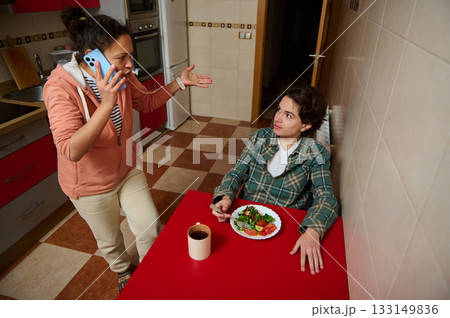 Teenage Boy With Salad At Red Table While Woman On Phone Argues In Kitchen Home Life Teenage Boy With Salad At Red Table While Woman On Phone Argues In Kitchen Home Life 133149836