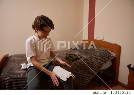 Teenage Boy Sitting On Bed, Smiling While Unfolding Towel In Cozy Bedroom 133150159