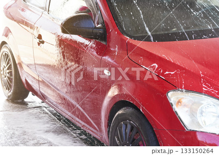 Red car washed in self serve carwash, detail on side covered with water sprayed from high pressure stream hose 133150264
