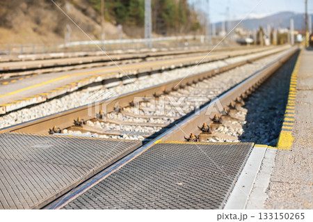 Empty train station in small city on sunny day, shallow depth of field photo, focus on concrete platform, passenger crossing, and steel rail foreground Empty train station in small city on sunny day, shallow depth of field photo, focus on concrete platform, passenger crossing, and steel rail foreground 133150265