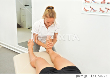 Young female physiotherapist massaging / exercising muscles at ankle and feet of her patient lying near Young female physiotherapist massaging / exercising muscles at ankle and feet of her patient lying near 133150277
