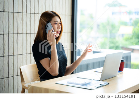 Asian businesswoman on smartphone while reviewing clipboard and laptop in a modern remote office. 133154191