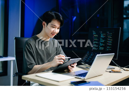 Young male programmer working with tablet and screen displaying code. Modern tech office setup, great for software, coding, IT 133154256
