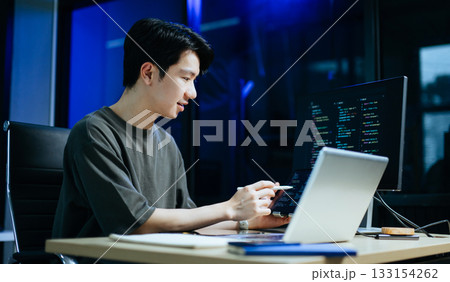 Young male programmer working with tablet and screen displaying code. Modern tech office setup, great for software, coding, IT 133154262