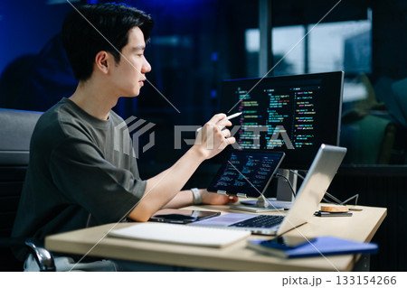 Young male programmer working with tablet and screen displaying code. Modern tech office setup, great for software, coding, IT 133154266