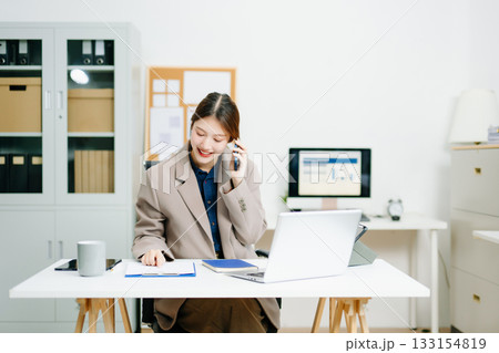 Smiling businesswoman talking on phone while working with laptop in a modern office. Perfect for themes of communication, marketing, finance Smiling businesswoman talking on phone while working with laptop in a modern office. Perfect for themes of communication, marketing, finance 133154819
