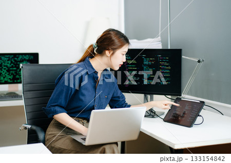 Young female programmer working with tablet and screen displaying code. Modern tech office setup, great for software, coding, IT Young female programmer working with tablet and screen displaying code. Modern tech office setup, great for software, coding, IT 133154842