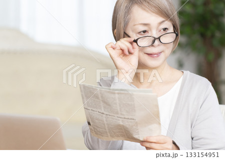 リビングで老眼鏡をかけて新聞を読むシニア女性 リビングで老眼鏡をかけて新聞を読むシニア女性 133154951