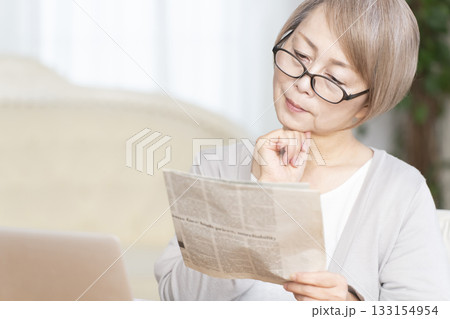 リビングで老眼鏡をかけて新聞を読むシニア女性 リビングで老眼鏡をかけて新聞を読むシニア女性 133154954