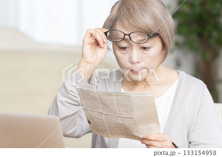 リビングで老眼鏡をかけて新聞を読むシニア女性 リビングで老眼鏡をかけて新聞を読むシニア女性 133154958