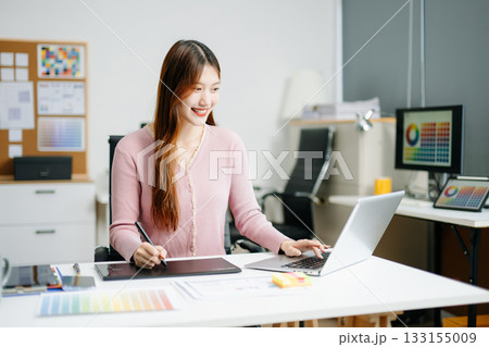 Young female graphic designer using digital tablet and laptop in modern office. Creative workspace, color palettes, and UI UX design 133155009