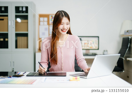 Young female graphic designer using digital tablet and laptop in modern office. Creative workspace, color palettes, and UI UX design 133155010