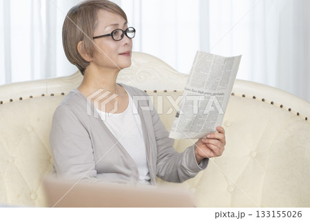 リビングのソファーに座り老眼鏡をかけて新聞を読むシニア女性 133155026