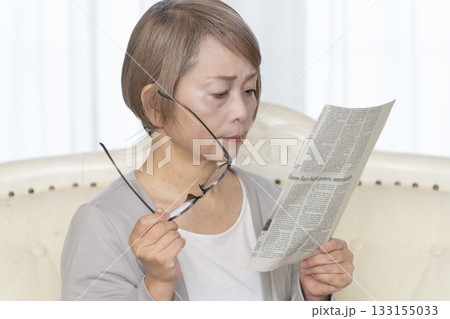リビングのソファーに座り老眼鏡をかけて新聞を読むシニア女性 リビングのソファーに座り老眼鏡をかけて新聞を読むシニア女性 133155033
