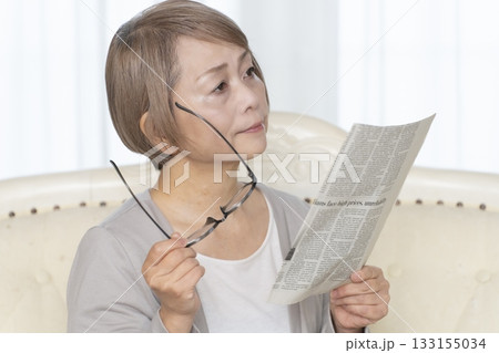 リビングのソファーに座り老眼鏡をかけて新聞を読むシニア女性 リビングのソファーに座り老眼鏡をかけて新聞を読むシニア女性 133155034