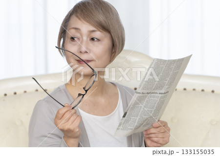 リビングのソファーに座り老眼鏡をかけて新聞を読むシニア女性 133155035