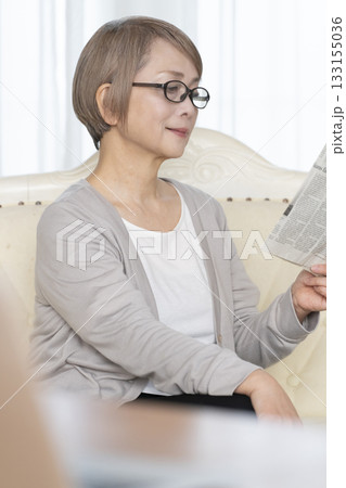リビングのソファーに座り老眼鏡をかけて新聞を読むシニア女性 リビングのソファーに座り老眼鏡をかけて新聞を読むシニア女性 133155036