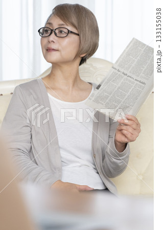 リビングのソファーに座り老眼鏡をかけて新聞を読むシニア女性 リビングのソファーに座り老眼鏡をかけて新聞を読むシニア女性 133155038