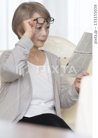 リビングのソファーに座り老眼鏡をかけて新聞を読むシニア女性 133155039
