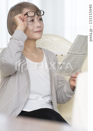 リビングのソファーに座り老眼鏡をかけて新聞を読むシニア女性 133155040