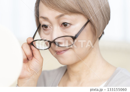 リビングで老眼鏡をかけて手鏡を見るシニア女性 リビングで老眼鏡をかけて手鏡を見るシニア女性 133155060