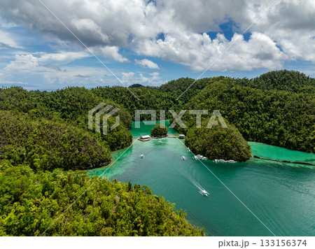 White boats sail through turquoise lagoon waters framed by forested hills. Siargao, Philippines. Sugba Blue Lagoon. 133156374