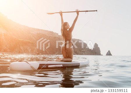 Woman Paddleboard Ocean - A woman celebrates after successfully paddleboarding on a calm ocean with cliffs in the background. 133159511