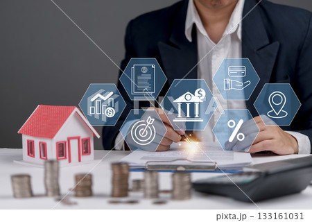 businessman in a suit working on a desk with a model house and icons related to real estate, mortgage, and financial planning. Property in 133161031