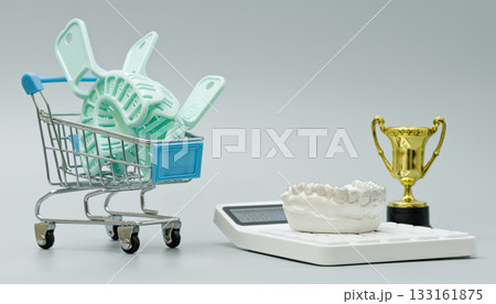 Mini shopping cart filled with dental impression trays beside a jaw model, calculator, and trophy, symbolizing cost, quality, and dental excellence. 133161875