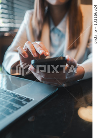 Close-up of a businesswoman's hands using a smartphone at a desk with a laptop, showcasing modern technology in a professional environment. Close-up of a businesswoman's hands using a smartphone at a desk with a laptop, showcasing modern technology in a professional environment. 133166924