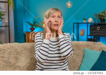 Mature woman looking upset anxious while listening to bad news on mobile phone sitting on home sofa Mature woman looking upset anxious while listening to bad news on mobile phone sitting on home sofa 133167441