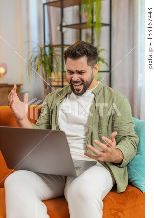 Upset young Caucasian man freelancer using laptop engages in heated conversations sitting on sofa 133167443