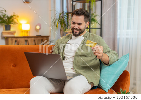Happy young man using credit bank card and laptop while shopping online sitting on sofa couch 133167588