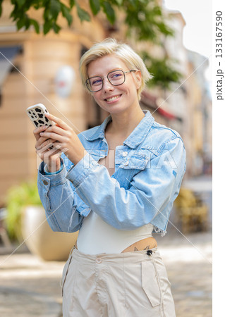 Smiling blond woman using smartphone typing texting scrolling social media app on city town street 133167590