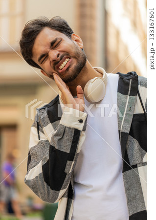 Upset Hispanic adult young man traveler feels a toothache while standing in urban city street 133167601
