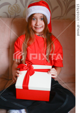 A girl wearing a red blouse and a Santa Claus hat opens a New Year's gift with a bow. The girl is happily surprised A girl wearing a red blouse and a Santa Claus hat opens a New Year's gift with a bow. The girl is happily surprised 133167615