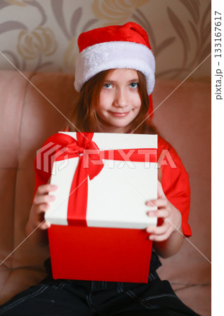 A girl wearing a red blouse and a Santa Claus hat opens a New Year's gift with a bow. The girl is happily surprised 133167617
